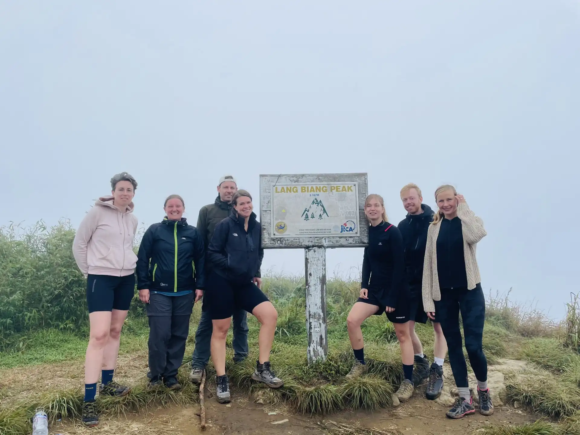 Trekking group celebrating at Lang Biang Peak 2167m summit sign Dalat Seven international hikers standing together at the Lang Biang Peak 2167M wooden elevation signboard in Dalat